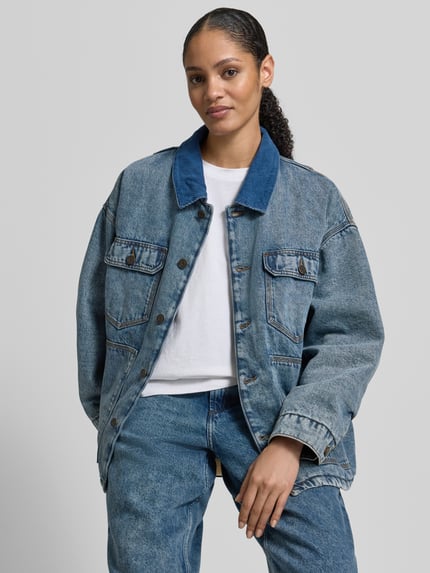 Vrouwenmodel draagt Jeansjacke mit Knopfleiste van American Vintage, losse pasvorm, blauwe denim, borst- en steekzakken, voorkant, bovenlichaam close-up.