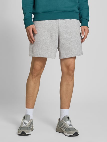 Männermodel trägt Relaxed Fit Shorts mit Logo-Stitching von New Balance in Hellgrau, Baumwoll-Mix, elastischer Bund, Ganzkörperansicht von vorne.