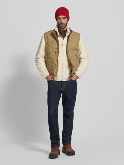 Männermodel trägt Weste mit Stehkragen von Polo Ralph Lauren in Beige, kombiniert mit Jeans und Wolljacke; Ganzkörperansicht von vorne.