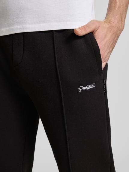 Mężczyznamodel nosi Loose Fit Sweatpants mit Label-Stitching und elastischem Bund von PROHIBITED, mieszanka bawełny, czarne spodnie z białym haftowanym logo, długość wewnętrzna nogawki w rozmiarze M: 80 cm, zbliżenie na górną część nogawek