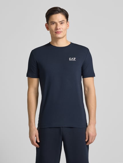 Männermodel trägt T-Shirt mit Label-Print von EA7 Emporio Armani in Dunkelblau, Slim Fit aus Baumwoll-Elasthan, Ganzkörperansicht von vorne.