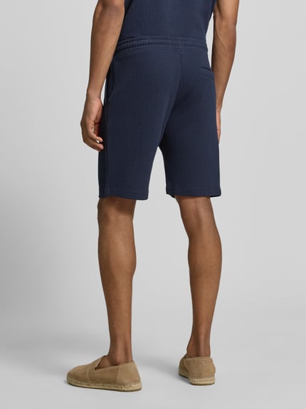 Männermodel trägt Regular Fit Shorts mit Tunnelzug von MCNEAL, mieszanka bawełny z dodatkiem streczu, krój regular fit, wpuszczane kieszenie, kieszeń z tyłu, szerokość w rozmiarze M 43 cm, długość wewnętrzna nogawki 23 cm, kolor granatowy, tekstura prążkowana, widok całej sylwetki z tyłu.