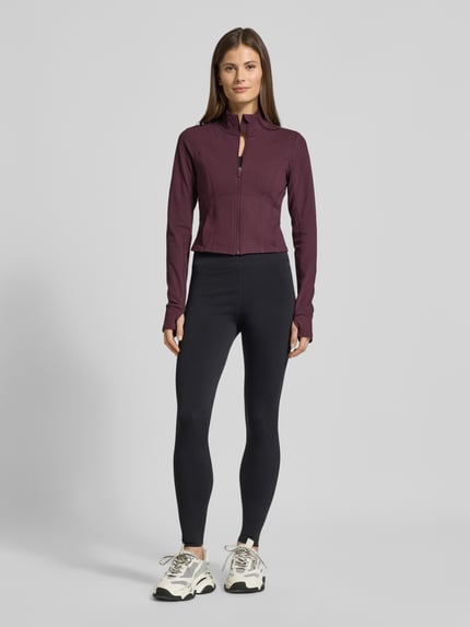Kobiece modelka nosi kurtkę Slim Fit Jacke mit Stehkragen Modell 'Storm' od Fabletics w kolorze ciemnofioletowym, gładka faktura, pętelki na kciuki, krój slim fit, krótki krój, Szerokość ramion w rozmiarze S: 36 cm, Szerokość klatki piersiowej w rozmiarze S: 42 cm, Długość tyłu w rozmiarze S: 44 cm, Długość rękawów w rozmiarze S: 74 cm, widok całej sylwetki z przodu.