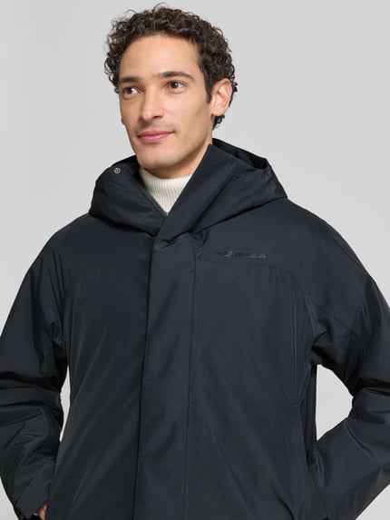 Męski model nosi czarną parkę Peak Performance Regular Fit Parka mit Kapuze Model 'Treeline HIPE', z kapturem, zamek błyskawiczny zakryty, Długość tyłu w rozmiarze M: 99 cm, Szerokość ramion w rozmiarze M: 54 cm, Szerokość w rozmiarze M: 60 cm, Długość rękawów w rozmiarze M: 61 cm, widok na górną część ciała z przodu.
