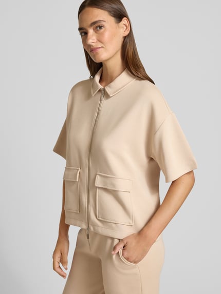 Modelka nosi 'Jacke mit Umlegekragen' od Christian Berg Woman, beżowy elastyczny materiał, proste cięcie, krótkie rękawy, kieszenie, widok z przodu na całą sylwetkę.