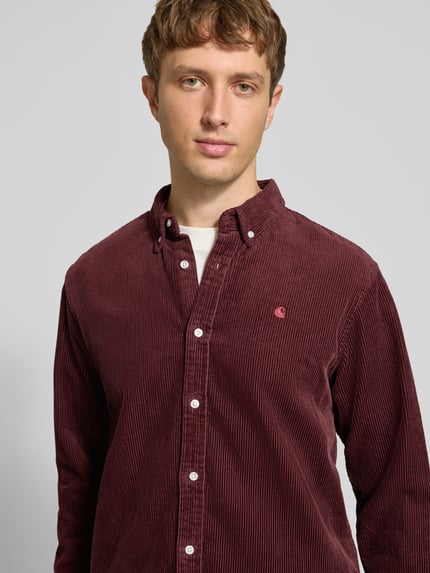 Mężczyzna nosi Regular Fit Freizeithemd mit Logo-Stitching Modell 'MADISON' od Carhartt Work In Progress, bordowy, bawełniany, z kołnierzykiem button-down, z przodu haftowane logo, zapięcie na guziki, teksturowana powierzchnia, krój regular fit, widok na górną część ciała z przodu.