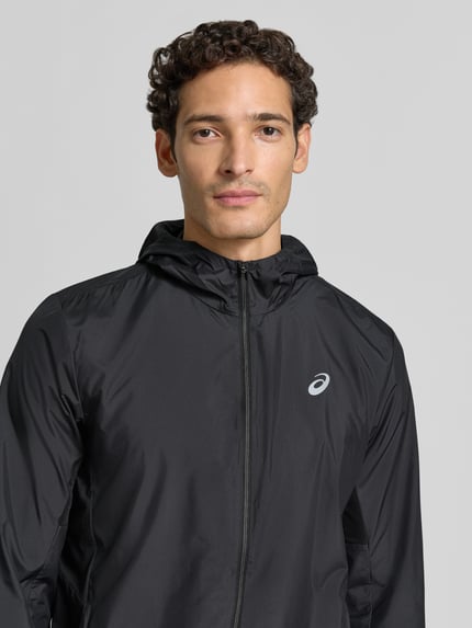 Männermodel trägt schwarze Regular Fit Trainingsjacke mit Logo-Print Modell 'Core' von ASICS TIGER, glatte Textur, Nahaufnahme Oberkörper.