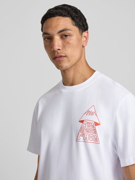 Männermodel trägt weißes Herren T-Shirt mit Motiv-Print Modell 'MOUNTAIN PLAY' von The North Face aus reiner Baumwolle, Regular-Fit, Rundhalsausschnitt, vorderseitigem Motiv-Print und rückseitigem Logo-Print, Nahaufnahme Oberkörper.