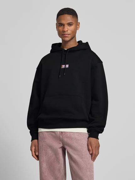 Männermodel trägt Hoodie mit Logo-Print Modell 'Chase' von Les Deux in zwart, loose fit, katoen, kangoeroezak, voorkant, Nahaufnahme Oberkörper.