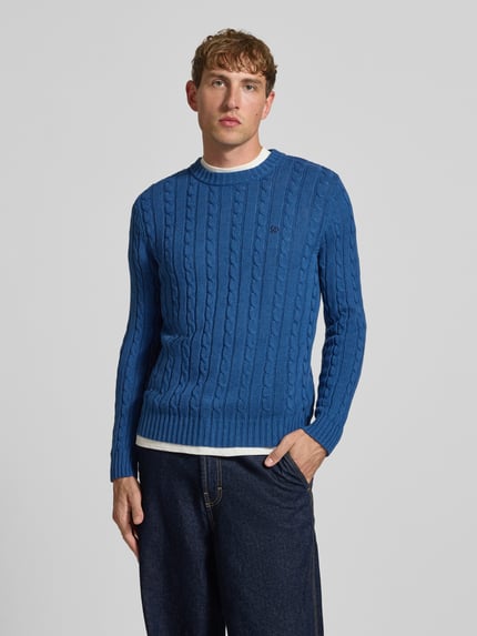 Männermodel trägt Strickpullover mit Zopfmuster und Rundhalsausschnitt von Superdry in Blau, reine Baumwolle, Slim Fit, Ganzkörperansicht von vorne.