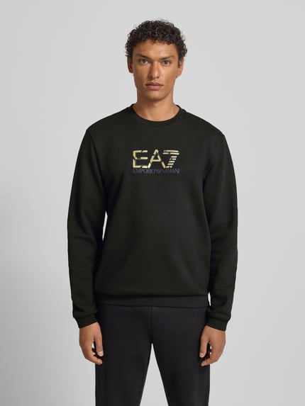 Męski model nosi bluzę Sweatshirt mit Logo und Rundhalsausschnitt od EA7 Emporio Armani, czarna bawełna, krój regular fit, logo, okrągły dekolt, długość tyłu 69 cm, długość rękawów 68 cm, całkowity widok z przodu.
