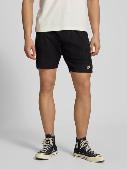 Männermodel trägt Shorts mit Label-Patch Modell 'LIBCO' von Pegador, Schwarz, Strukturmuster, Material-Mix, Gerader-Schnitt, Elastischer-Bund, Ganzkörperansicht von vorne.