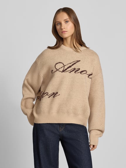 Frauenmodel trägt Oversized Strickpullover mit Label-Details von ANOTHER COTTON LAB, beige, weiche Struktur, gerade Schnittform, Nahaufnahme Oberkörper.