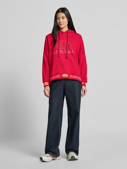 Vrouwenmodel draagt Oversized Hoodie uit puur katoen Model 'Namasté' van miss goodlife in rood met statementprint en met strass-steentjes, elastische boorden, frontale lichaamsopname.