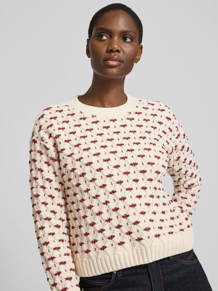 Frauenmodel trägt Pullover mit Strukturstrick von VERO MODA in Creme mit rot-lilafarbenem Blümchenmuster, Strickoptik, Nahaufnahme Oberkörper.