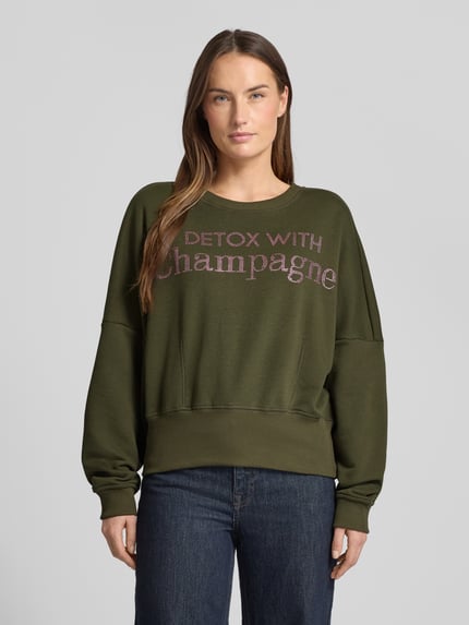 Vrouwenmodel draagt Oversized Sweatshirt uit pure katoen van miss goodlife, olijfgroen met statement-print, ronde hals, volledig vooraanzicht.