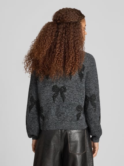 Frauenmodel trägt Pullover mit Glitzer-Schleifenprint von ONLY in melierter Strickoptik, Motiv-Print, Effektgarn, Ganzkörperansicht Rückansicht.