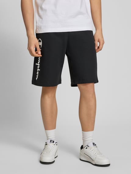 Männermodel trägt schwarze Shorts mit Logo-Print und -Stitching von CHAMPION, Baumwoll-Mix, lockerer Schnitt, Nahaufnahme Unterkörper.