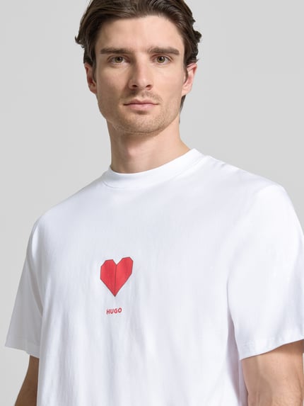 Männermodel trägt Regular Fit T-Shirt aus reiner Baumwolle Modell 'DALENTIN' von HUGO in Weiß mit rotem Herz-Print und Label, Nahaufnahme Oberkörper.