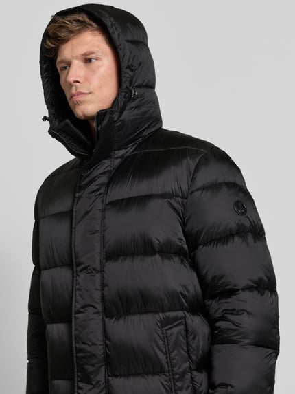 Männermodel trägt Pufferjacke mit Kapuze Modell 'JC-315Norman' von JOOP! in Schwarz mit Logo-Patch und Reißverschluss, Nahaufnahme Oberkörper.