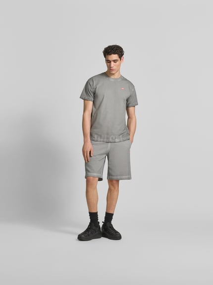 Męski model trägt Sweatshorts aus reiner Baumwolle von Diesel, luźny krój, elastyczny pas, efekt znoszenia, nadruk z logo, szary kolor, szerokość w rozmiarze M 38 cm, długość wewnętrzna nogawki 25 cm, Ganzkörperansicht von vorne.