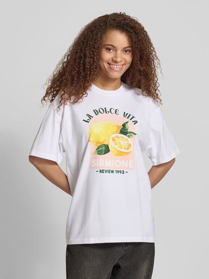 Kobiece modelka nosi damski t-shirt 'T-Shirt mit rückseitigem Motiv-Print' od REVIEW z bawełny, krój oversized, okrągły dekolt, długość tyłu 68 cm w rozmiarze S, widok z przodu, ujęcie całej sylwetki.