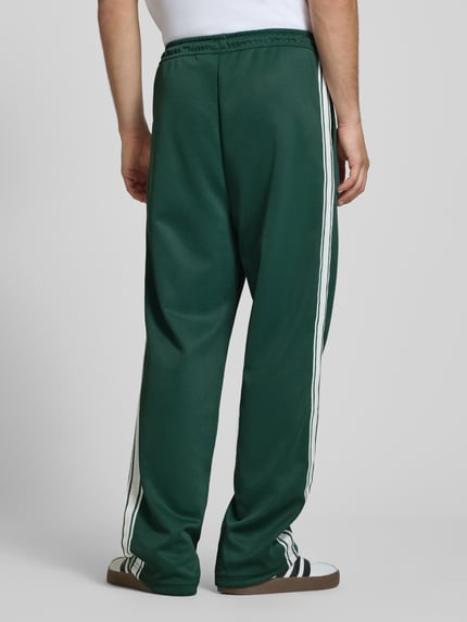 Männermodel trägt Wide Leg Sweatpants mit elastischem Bund von Pegador in Dunkelgrün mit weißen Seitenstreifen, Ganzkörperansicht Rückansicht.