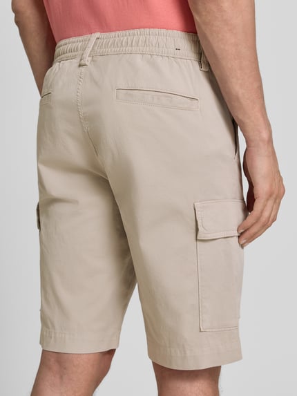 Männermodel trägt Relaxed Fit Cargoshorts aus Baumwoll-Mix Modell 'SISLA' von BOSS Orange, Optik: Unifarben, Passform: Relaxed-Fit, Label-Patch, seitliche Eingrifftaschen, Gesäßtaschen, Cargotaschen, Farbe Beige, Material Baumwoll-Mix, Ganzkörperansicht Rückansicht.