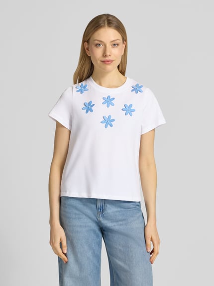 Frauenmodel trägt T-Shirt mit Stitchings und Perlen von Rich & Royal, weiße Reine Baumwolle, Regular-Fit, Rundhalsausschnitt, Rückenlänge bei Größe S 54 cm, blaue Stickerei als Blumenmotiv, lässige T-Shirt-Optik, Ganzkörperansicht von vorne.