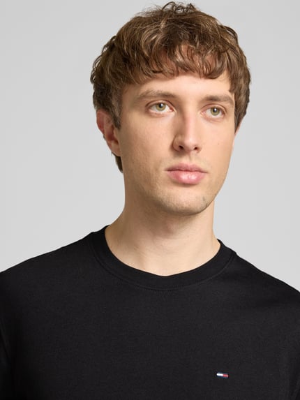 Mannelijk model draagt Regular Fit T-Shirt aus reiner Baumwolle van Tommy Jeans, zwarte unilook, logo-stitching, ronde hals, korte mouw, jersey puur katoen, Nahaufnahme bovenlichaam.