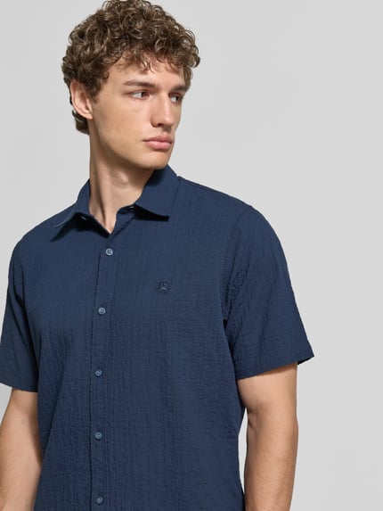 Mężczyznęmodel nosi Relaxed Fit Freizeithemd mit Logo-Stitching und Kentkragen von Lerros, kolor granatowy, faktura seersucker, listwa guzikowa, wyhaftowane logo, zaokrąglony dół, długość tyłu w rozmiarze M: 75 cm, szerokość ramion w rozmiarze M: 45 cm, długość rękawów w rozmiarze M: 25 cm, zbliżenie na górną część ciała