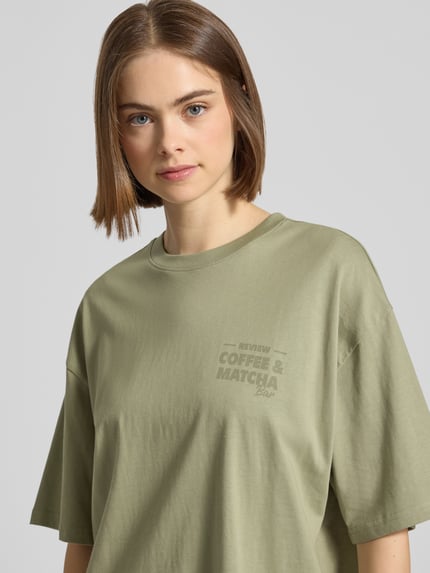 Vrouwenmodel draagt Oversized T-Shirt mit Statement-Print van REVIEW in olijfgroen katoen, ronde hals, oversized fit, detailopname bovenlichaam.