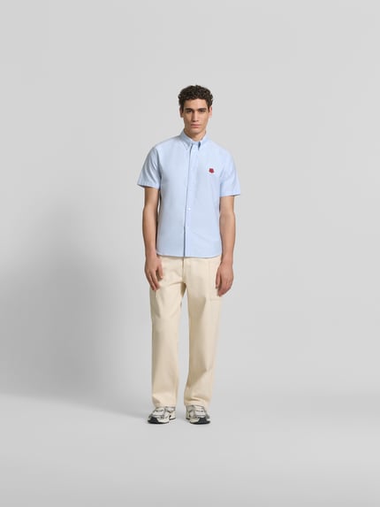 Męski model nosi Slim Fit Freizeithemd mit Logo-Stitching od Kenzo z bawełny, krój slim fit, kołnierzyk typu button down, krótki rękaw, jasnoniebieski, długość tyłu w rozmiarze M: 49 cm, szerokość w rozmiarze M: 77 cm, długość rękawów w rozmiarze M: 29 cm, haftowane logo, widok całej sylwetki z przodu.
