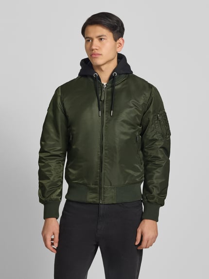 Männermodel trägt Bomberjacke mit Kapuze und Reißverschlusstaschen von Superdry in Olivgrün, Regular-Fit, Ganzkörperansicht von vorne, Rückenlänge bei Größe M 56 cm, Schulterbreite 45 cm, Brustweite 60 cm, Ärmellänge 68 cm.