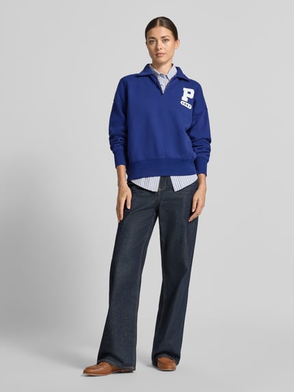 Frauenmodel trägt Loose Fit Sweatshirt aus Baumwoll-Mix von Polo Ralph Lauren in kräftigem Blau, kombiniert mit dunkelblauer Hose und Hemd, Ganzkörperansicht von vorne. Damen Sweatshirt von Polo Ralph Lauren.
