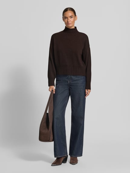 Kobiece model prezentuje sweter z dzianiny 'Strickpullover aus Woll-Mix mit Stehkragen' od Milano Italy, mieszanka wełny z domieszką kaszmiru, krój relaxed fit, stójka, opuszczone ramiona, długość tyłu w rozmiarze S: 51 cm, długość rękawa w rozmiarze S: 52 cm, ciemnobrązowa góra, klasyczny granatowy dół, tekstura miękka, gładkie tło, widok całej sylwetki z przodu.