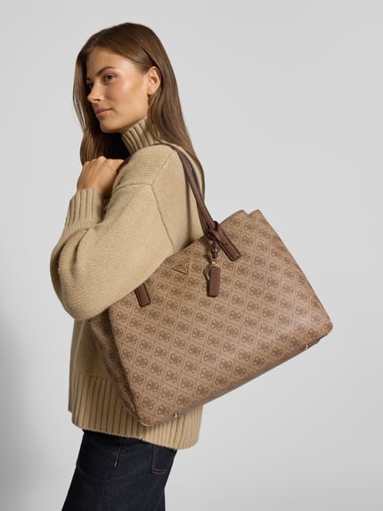 Frauenmodel trägt Handtasche in Leder-Optik Modell 'NOELLE' von GUESS mit Logo-Detail, beigebraun, Ganzkörperansicht von vorne. Geteiltes Hauptfach mit Magnet- und Reißverschluss, schmale Schulterriemen, Maße: 38x28x13 cm, Riemenlänge 64 cm.