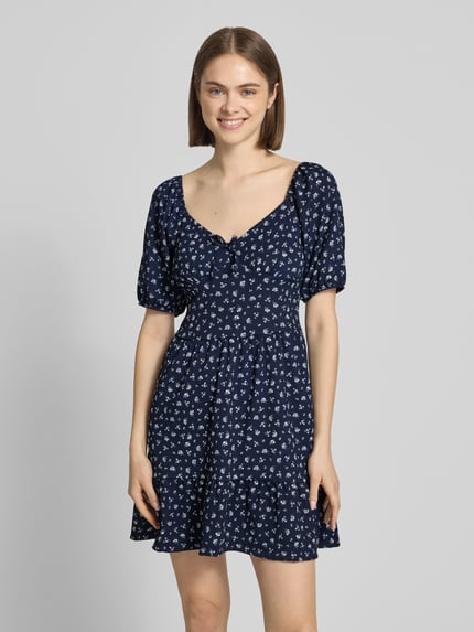 Vrouwenmodel draagt mini-jurk met pofmouwen model 'HILLARY' van Only, all-over bloemenmotief, crêpe materiaal, getailleerd, minilengte, vierkante hals, strikapplicatie. Ganzkörperansicht van voren.