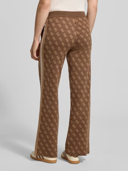 Frauenmodel trägt Wide Leg Strickhose mit Logo-Muster Modell 'ANEIRA' von GUESS, braun gemustert, elastischer Bund, Ganzkörperansicht Rückansicht. Material-Mix mit Woll-Anteil, Allover-Look.