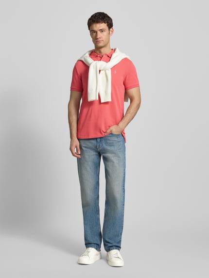 Męskimodel nosi Koszulkę polo z wyhaftowanym logo marki Polo Ralph Lauren z bawełny, krój regular fit, kołnierzyk polo, krótka listwa guzikowa, prążkowane zakończenia rękawów, długość tyłu w rozmiarze M: 68 cm, kolor różowy, tekstura gładka, jasnoniebieskie jeansy i białe sneakersy. Ganzkörperansicht von vorne.