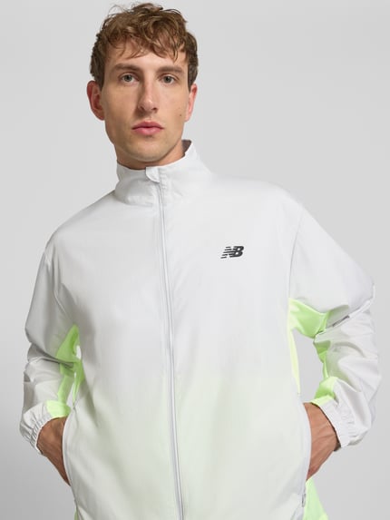 Männermodel trägt Regular Fit Jacke in Stehkragen Modell 'Fast Days' von New Balance in Weiß mit Logo-Print, Reißverschluss, Eingrifftaschen, elastischen Ärmelabschlüssen und Tunnelzug im Bund, Rückenlänge 68 cm, Ärmellänge 66 cm, Nahaufnahme Oberkörper.