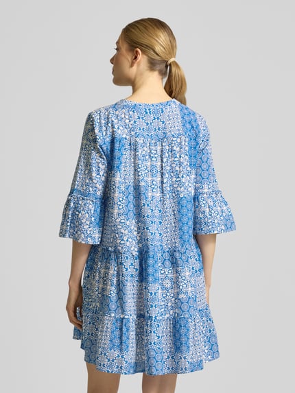 Frauenmodel trägt Ausgestelltes Blusenkleid aus reiner Baumwolle Modell 'DICTHE' von Vero Moda mit Optik:Allover-Muster, Mini-Länge, Raffung, Henley-Ausschnitt, 1/2-Arm, Trompetenärmel; farbenfrohes Muster in Blau- und Weißtönen, strukturierte leichte Textur, Ganzkörperansicht Rückansicht.