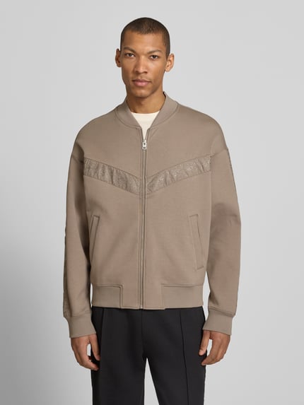 Männermodel trägt Oversized Sweatjacke aus Baumwoll-Mix Modell 'DROMZIP' von HUGO in Beige mit Reißverschluss und Logo-Patch, Ganzkörperansicht von vorne.