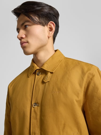 Männermodel trägt Jacke mit Label-Stitching und Eingrifftaschen von BLKVIS in Senfgelb, reine Baumwolle, Knopfleiste, Nahaufnahme Oberkörper.