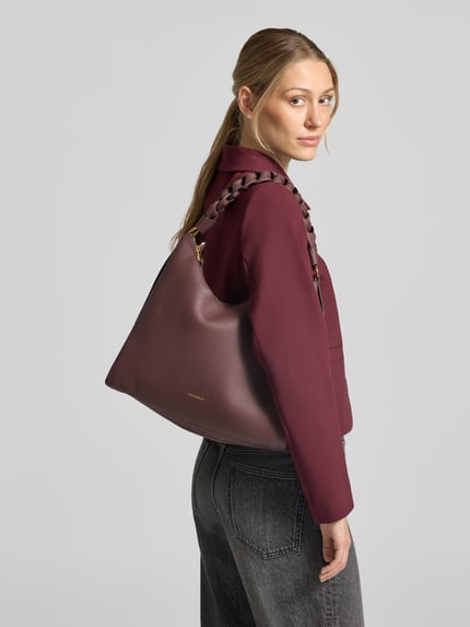 Frauenmodel trägt Handtasche aus echtem Leder Modell 'BOHEME' von Coccinelle in dunklem Rotbraun, glatte Textur, Ganzkörperansicht Rückansicht.