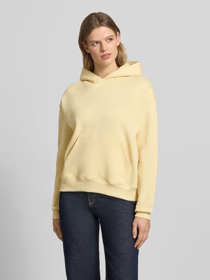 Kobiece model prezentuje Regular Fit Hoodie mit Känguru-Tasche Modell 'Iconic' von Gina Tricot, mieszanka bawełny, krój regular fit, kieszeń kangurka, elastyczne wykończenia, jasny kremowy kolor, przód, całopostaciowy widok.