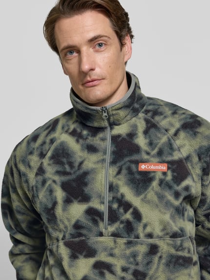 Männermodel trägt Fleecepullover mit Label-Details Modell 'Timber Crest™' von Columbia, grün-schwarzes Muster, Nahaufnahme Oberkörper. Gerader Schnitt, Stehkragen, Reißverschluss, Rückenlänge 70 cm, Ärmellänge 73 cm.
