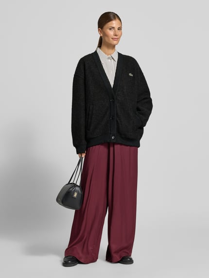 Frauenmodel trägt Cardigan mit Logo-Patch von Lacoste in Melange-Strickoptik, Strukturmuster, V-Ausschnitt, Knopfleiste, Ganzkörperansicht von vorne.