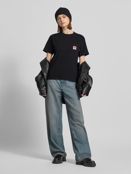 Frauenmodel trägt Damen T-Shirt mit Label-Patch von Carhartt Work In Progress in Schwarz, Reine Baumwolle, Gerader-Schnitt, Rundhalsausschnitt, Brusttasche, Rückenlänge 58 cm bei Größe S, Ganzkörperansicht von vorne.