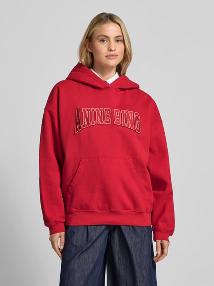 Vrouwenmodel draagt Oversized Hoodie uit puur katoen van Anine Bing in rood met labelprint en kangoeroezak, ribboorden, Ganzlichaamsvoorzijde. Ruglengte maat S: 65 cm.